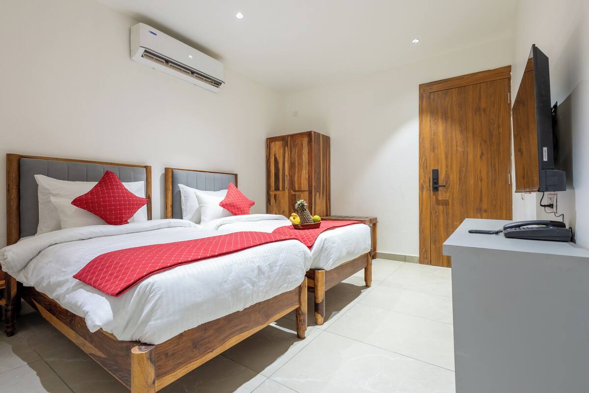 A double bed with red pillows, a wooden wardrobe, a desk with a telephone, and an air conditioning unit.