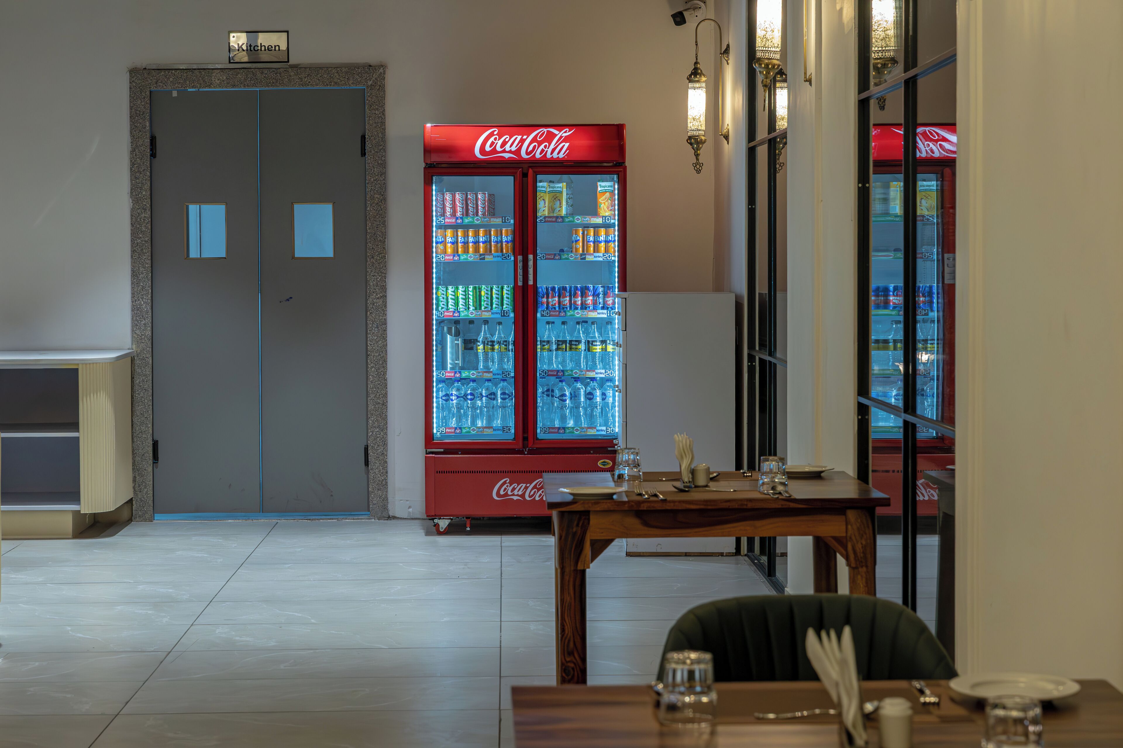 A dining area with a table set for two, a Coca-Cola refrigerator, and a kitchen door.