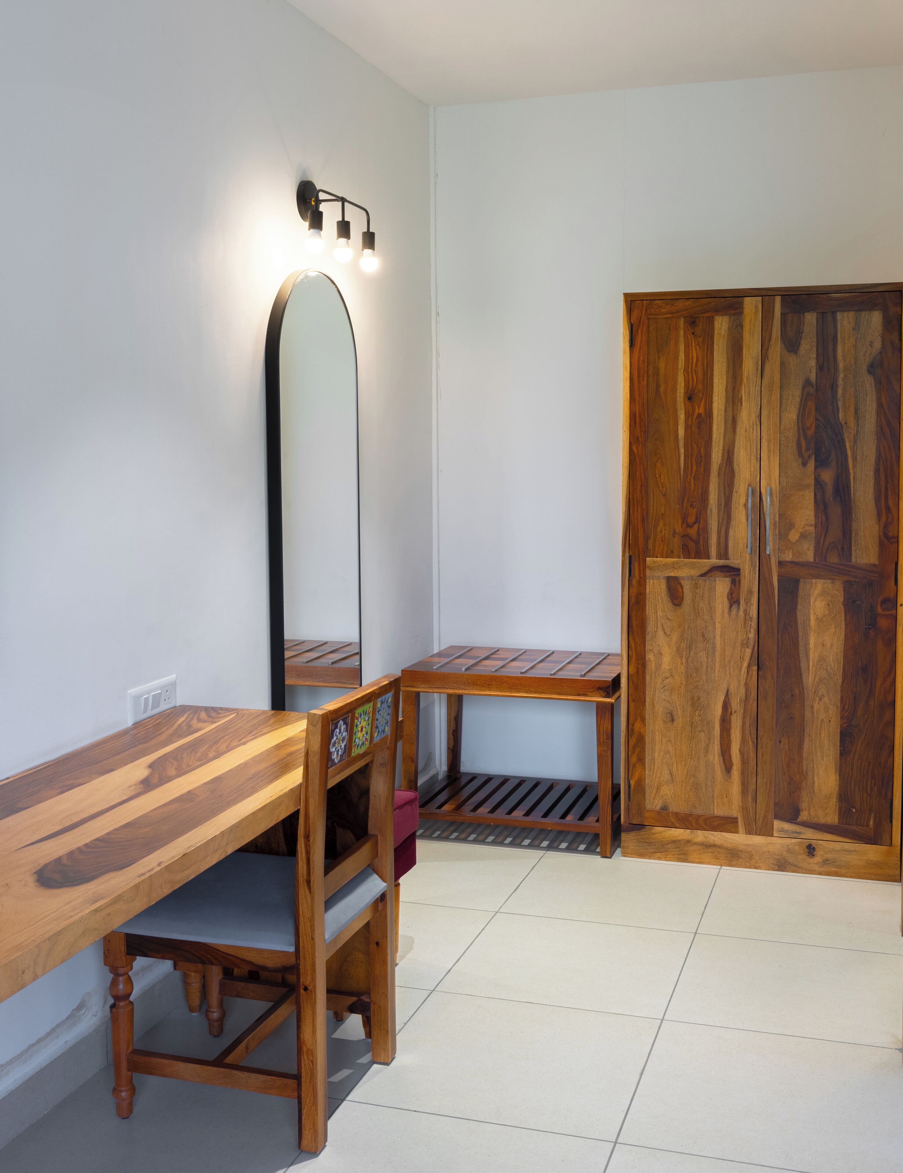 A room with a wooden desk, a mirror, a wardrobe, and a tiled floor.