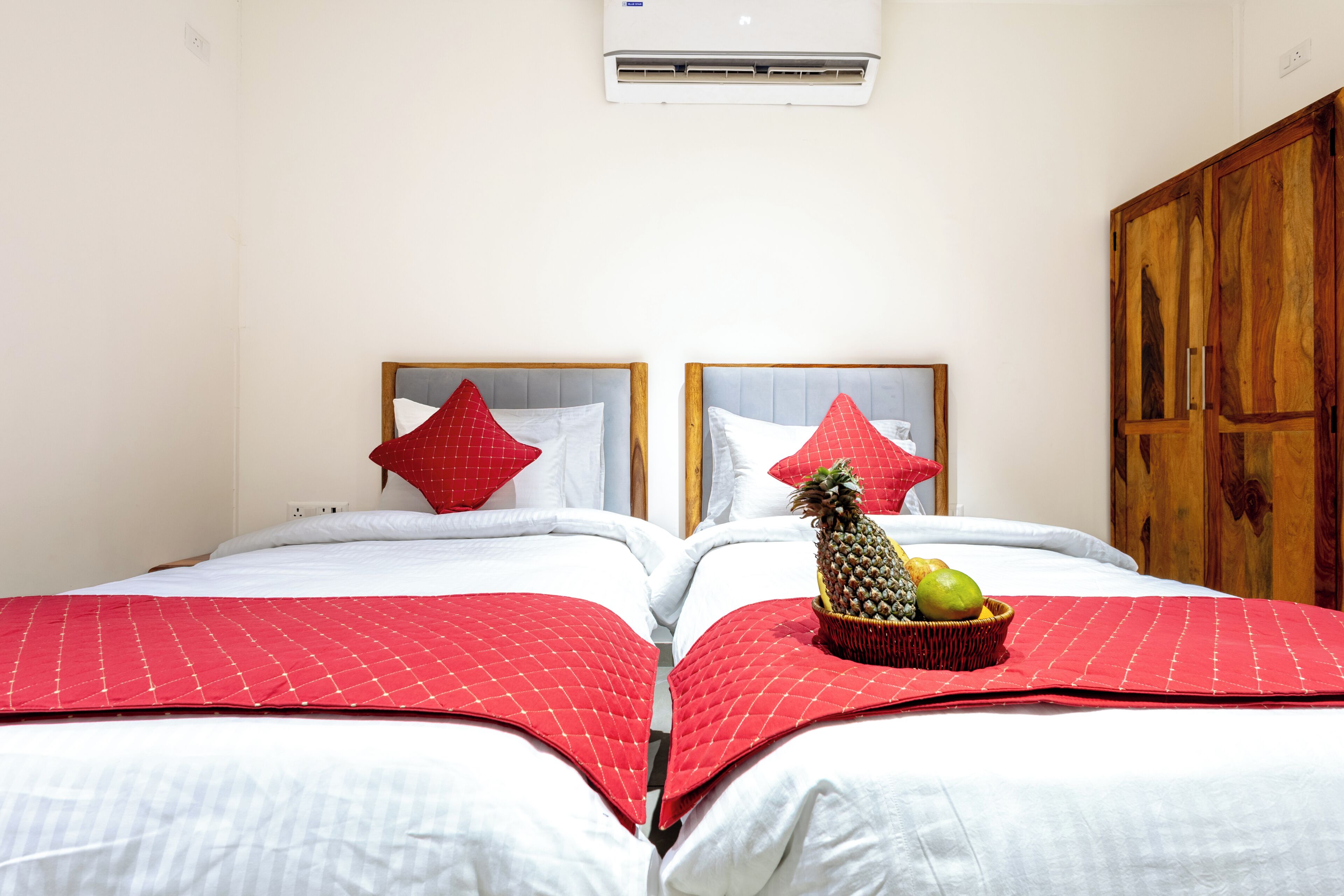 A hotel room with two beds, red and white bedding, a fruit basket on one bed, and a wooden wardrobe.