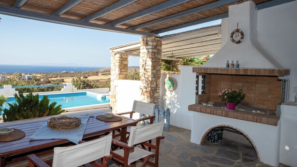 Un área de comedor al aire libre techada, con pilares de piedra, una piscina y vistas al mar.