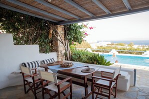 Ein Außenbereich mit Sitzgelegenheit, Blick auf einen Pool, Steinboden und einer Holzpergola.