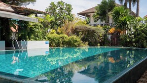 En rektangulär swimmingpool med klart vatten, omgiven av frodig grönska och en byggnad med ett tak täckt av tegelpannor.