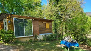 A wooden cabin with a large window, surrounded by greenery and a small outdoor seating area. - CHALET RENTAL IN PERIGORD NOIR (Prats-du-Périgord)