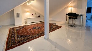 Une pièce mansardée avec un plafond blanc, un grand tapis à motifs, une console blanche agrémentée d’objets décoratifs, une chaise noire et un téléviseur fixé au mur.