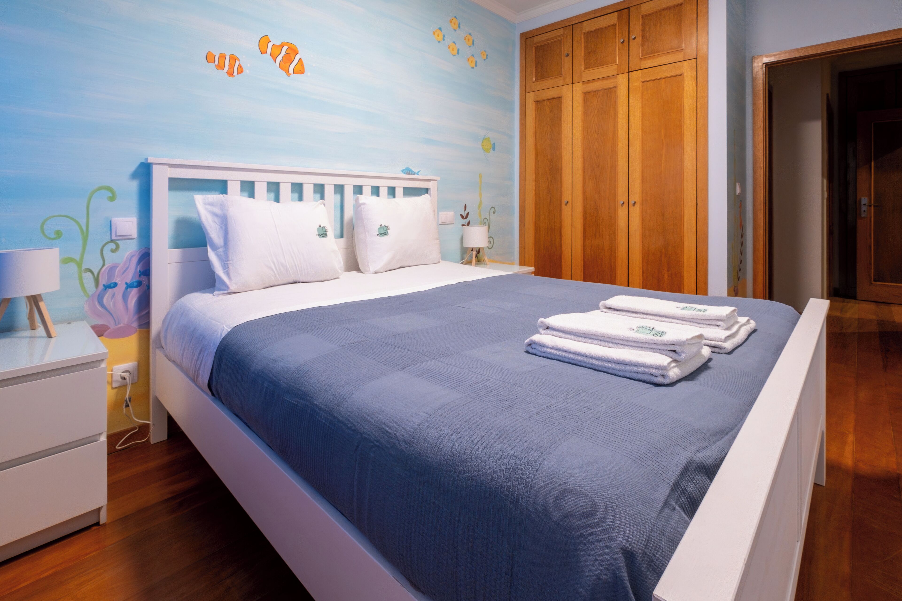 A bedroom with a bed, wooden wardrobe, and a wall with a blue and yellow mural.