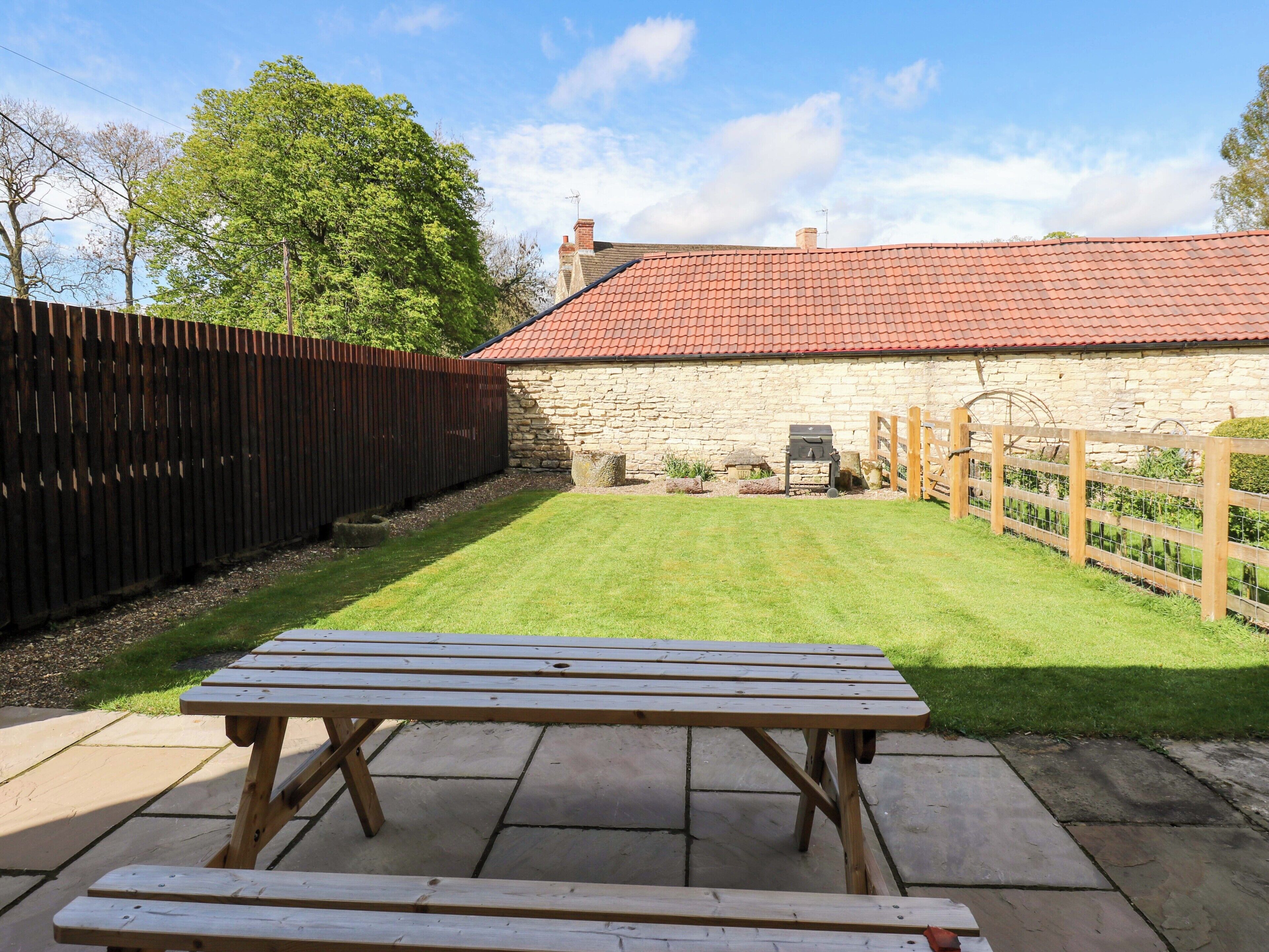 A stone building with a red-tiled roof, a wooden fence, and a picnic table in a grassy yard.
