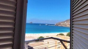 Une vue depuis une fenêtre donnant sur une plage, avec des bateaux, un ciel dégagé et des collines lointaines.