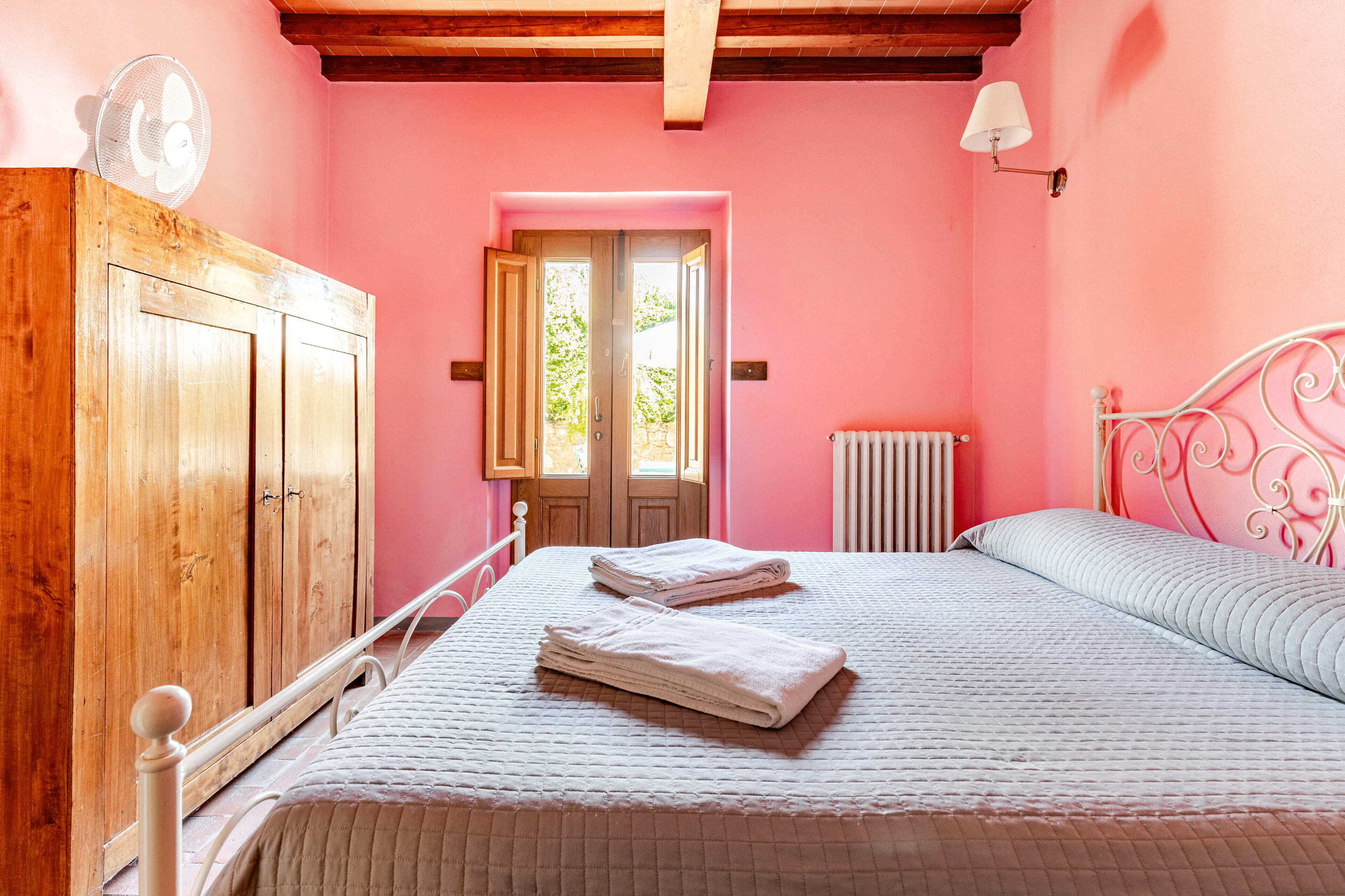 A bedroom with a wooden wardrobe, a bed with white bedding, a radiator, and a window with a view of greenery.