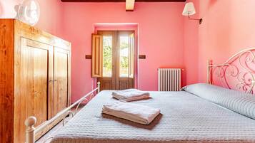 A bedroom with a wooden wardrobe, a bed with white bedding, a radiator, and a window with a view of greenery.