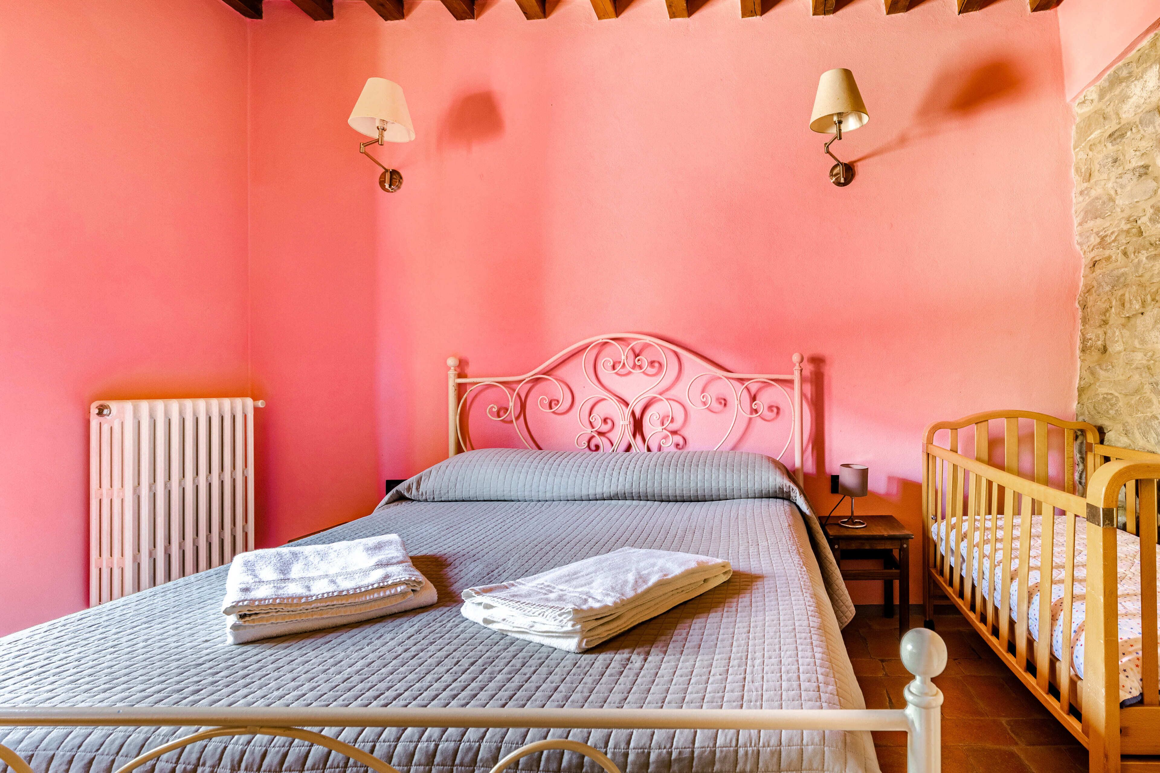 Camera da letto con parete rosa, letto con testiera in metallo, due lampade da comodino, una culla in legno e un termosifone.
