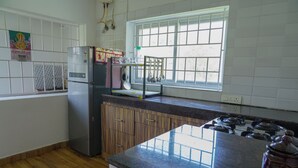 A kitchen with a refrigerator, stove, wooden cabinets, a window with blinds, and a hanging rack with kitchen utensils. - Winterbourne Chalet 3-bedroom villa in pleasant Coonoor getaway (Coonoor)