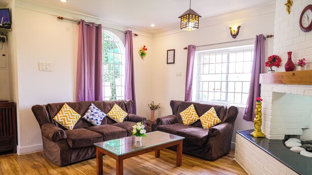 A living room with two brown sofas, a glass coffee table, wooden flooring, purple curtains, a fireplace, and a clock on the wall. - Winterbourne Chalet 3-bedroom villa in pleasant Coonoor getaway (Coonoor)