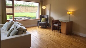 A room with a sofa, a chair, a desk, a lamp, and a window with a view of greenery. - Luxury 2 Bed Lodge, Beside Castlemartyr Resort (Castlemartyr)