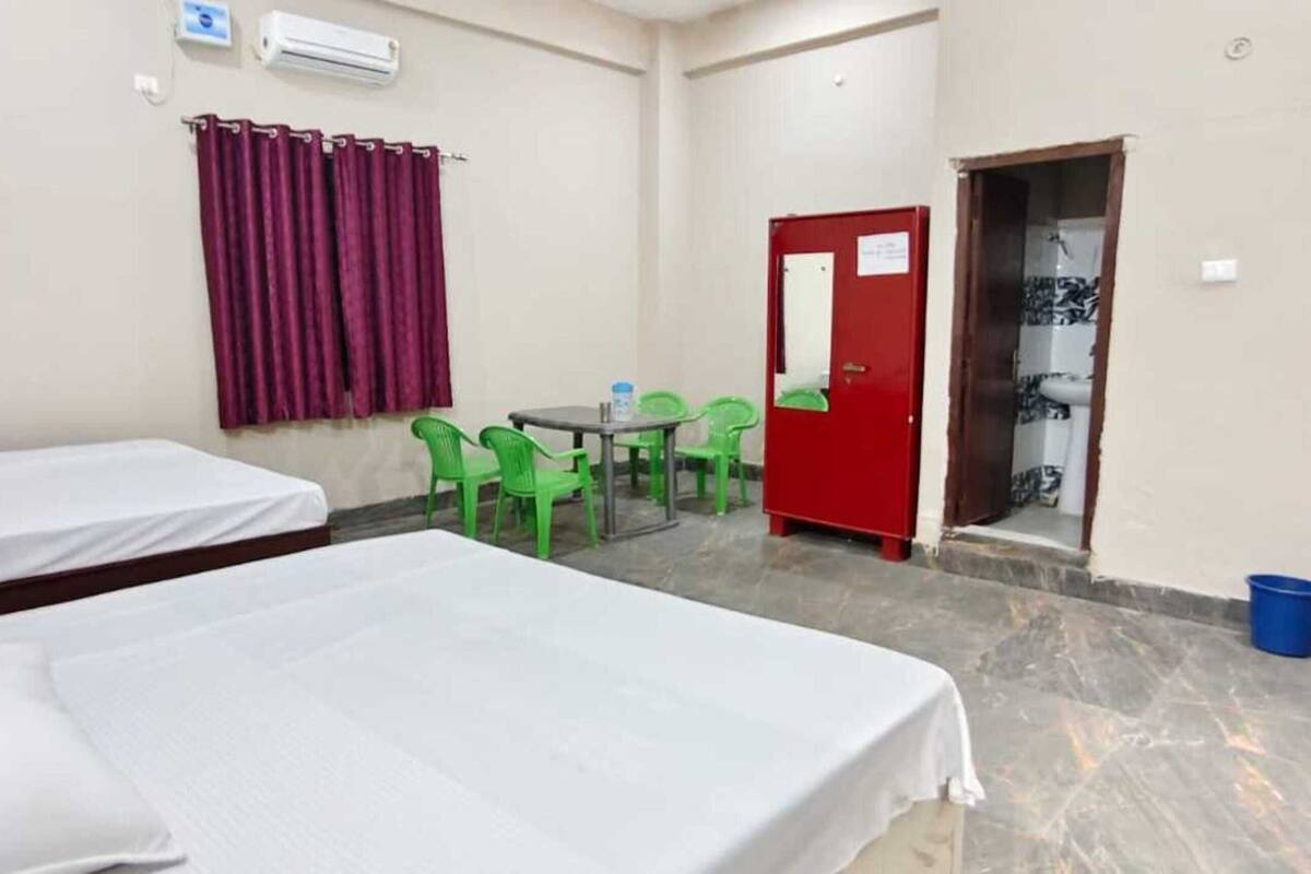 A hotel room with two beds, a table with chairs, a red door, and an air conditioning unit.