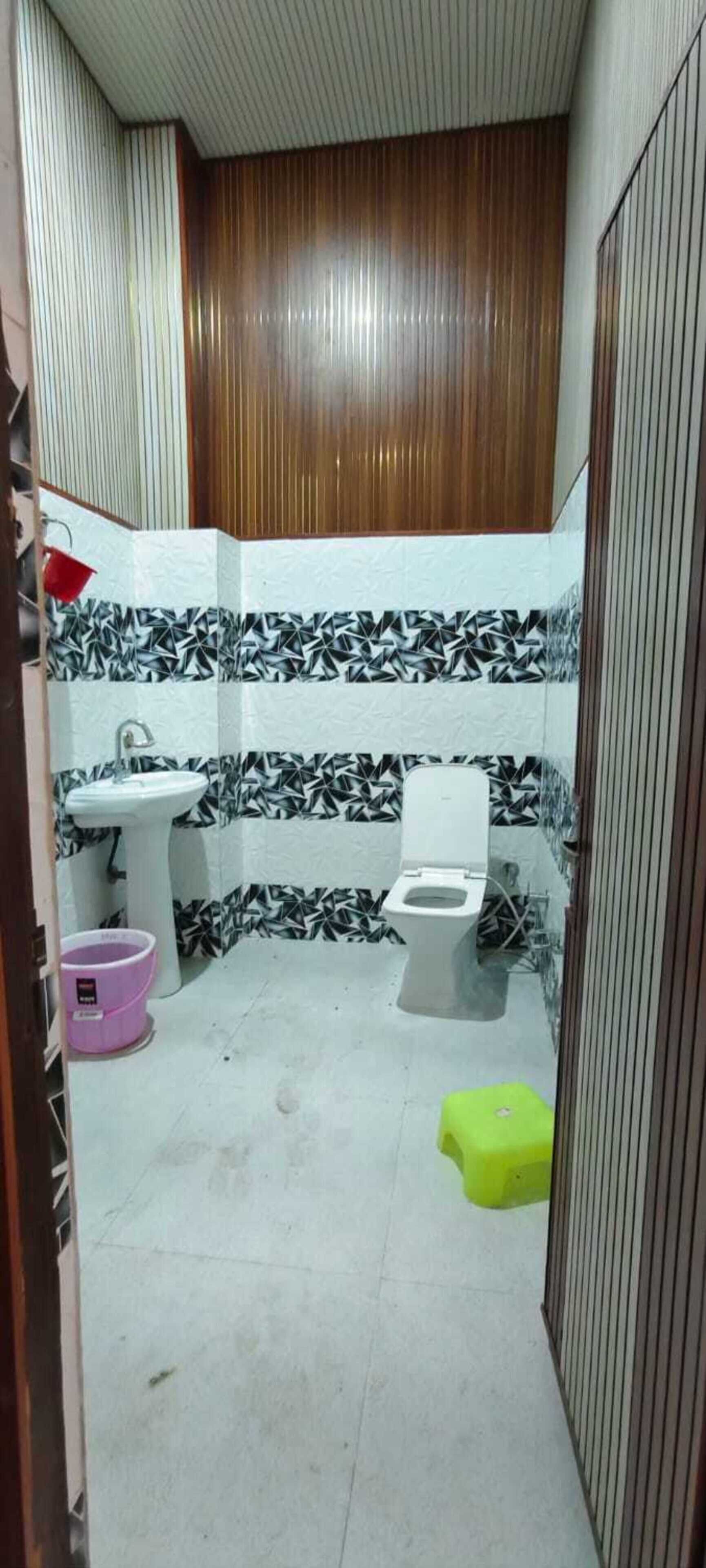 A small bathroom with a toilet, sink, and a mirror. There is a pink bucket and a green stool.