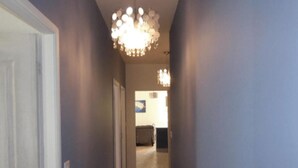 A hallway with a chandelier, a door on the left, and a kitchen area in the distance. - GITE HIBISCUS (Sanilhac)