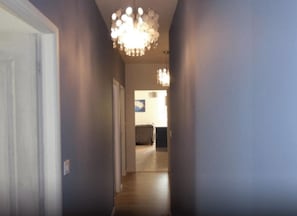 Ein Flur mit einem Chandelier, einer Tür links und einer weiter hinten liegenden Küchenzeile.