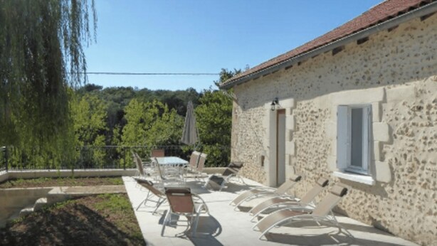 A stone building with a tiled roof, outdoor seating with umbrellas, and a view of trees and hills. - GITE HIBISCUS (Sanilhac)