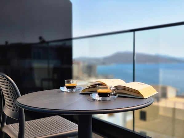 A round table with two cups of coffee and an open book, overlooking a body of water and buildings. - Rêve Studio Apartments (Sarande)