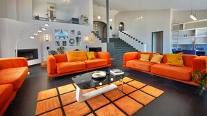 Ein geräumiges Wohnzimmer mit zwei orangefarbenen Sofas, einem Glastisch und einem gemusterten Teppich. Im Hintergrund sind ein Kamin und eine Treppe zu sehen.