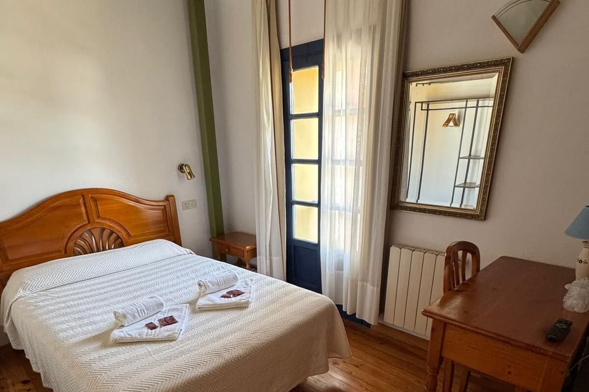 Una habitación con una cama, una mesa de madera, una silla, una ventana con cortinas y un espejo en la pared.