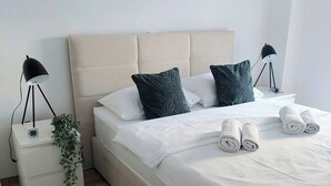 Un lit bien fait, avec du linge de lit blanc et des serviettes pliées, une tête de lit recouverte d’un tissu beige, deux lampes de chevet et une petite plante placée sur la table de chevet.