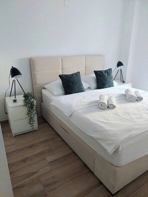 Ein ordentlich bezogenes Bett mit weißen Leinen und aufgerollten Handtüchern, ein beige farbener möbelbezogener Kopfteil, zwei Nachttischlampen und eine kleine Pflanze auf dem Nachttisch.