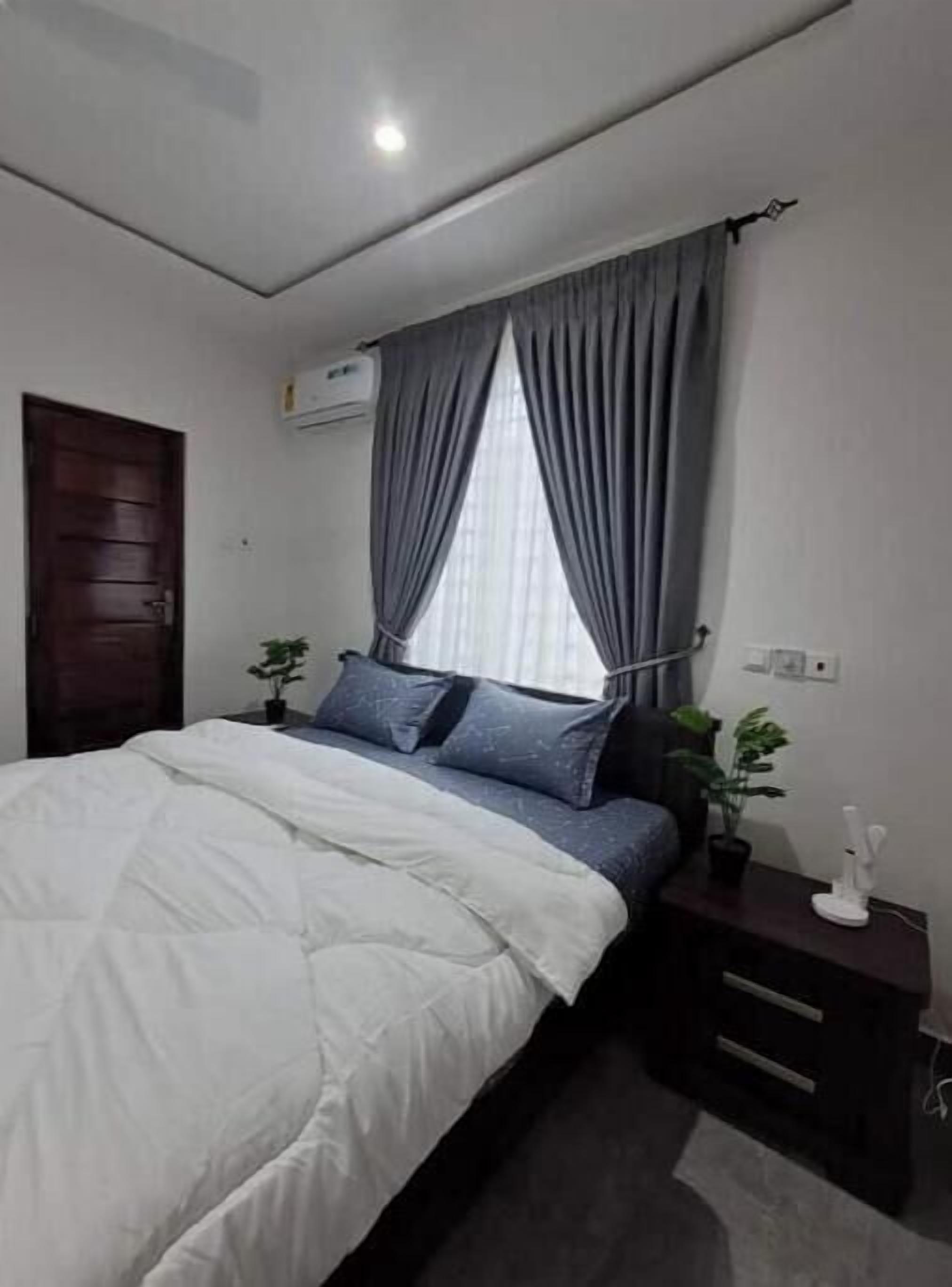 Un dormitorio con cama, mesita de noche, planta en maceta y ventana con cortinas.