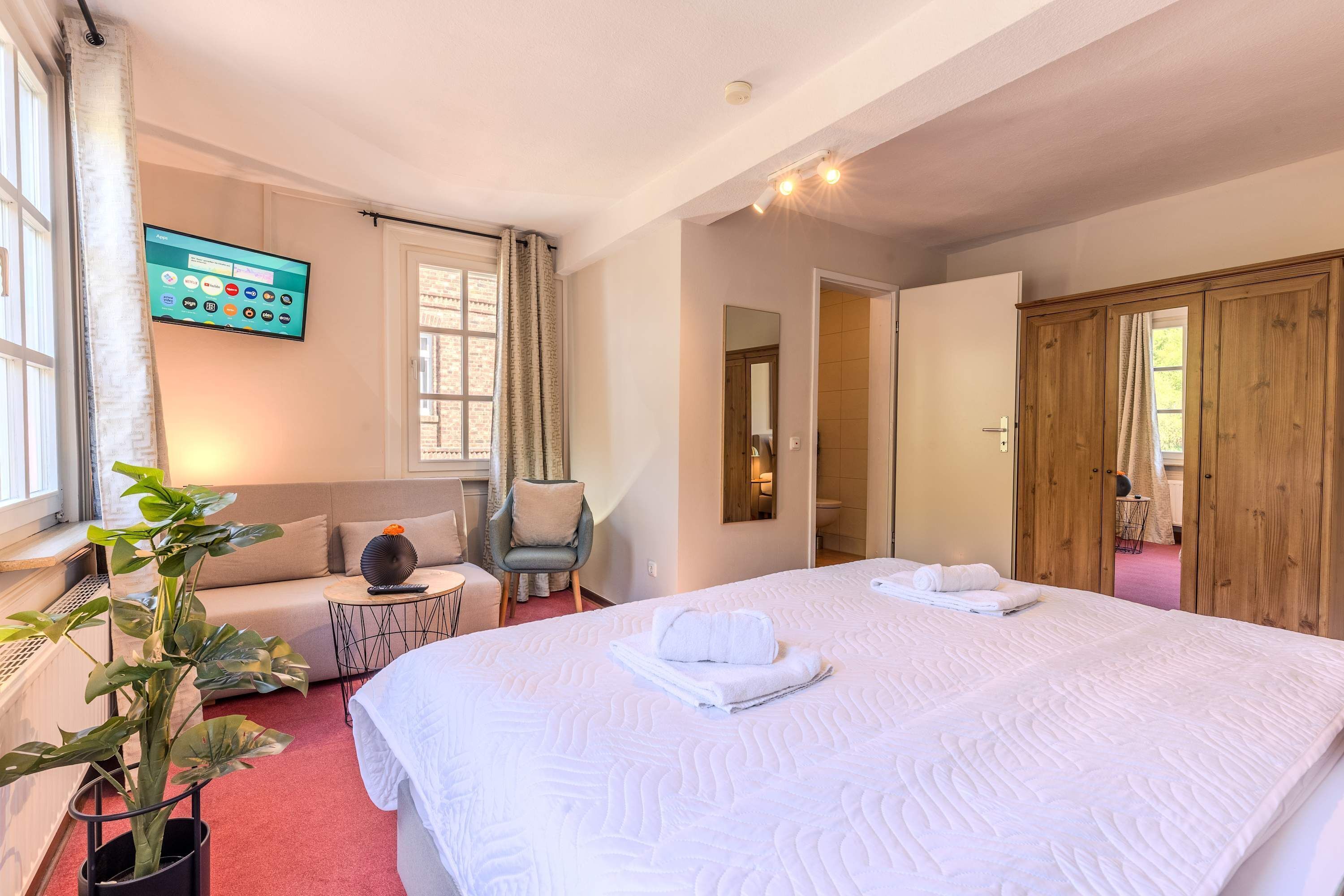 A hotel room with a large bed, a TV mounted on the wall, a potted plant, a seating area with a chair and table, and a wooden wardrobe.