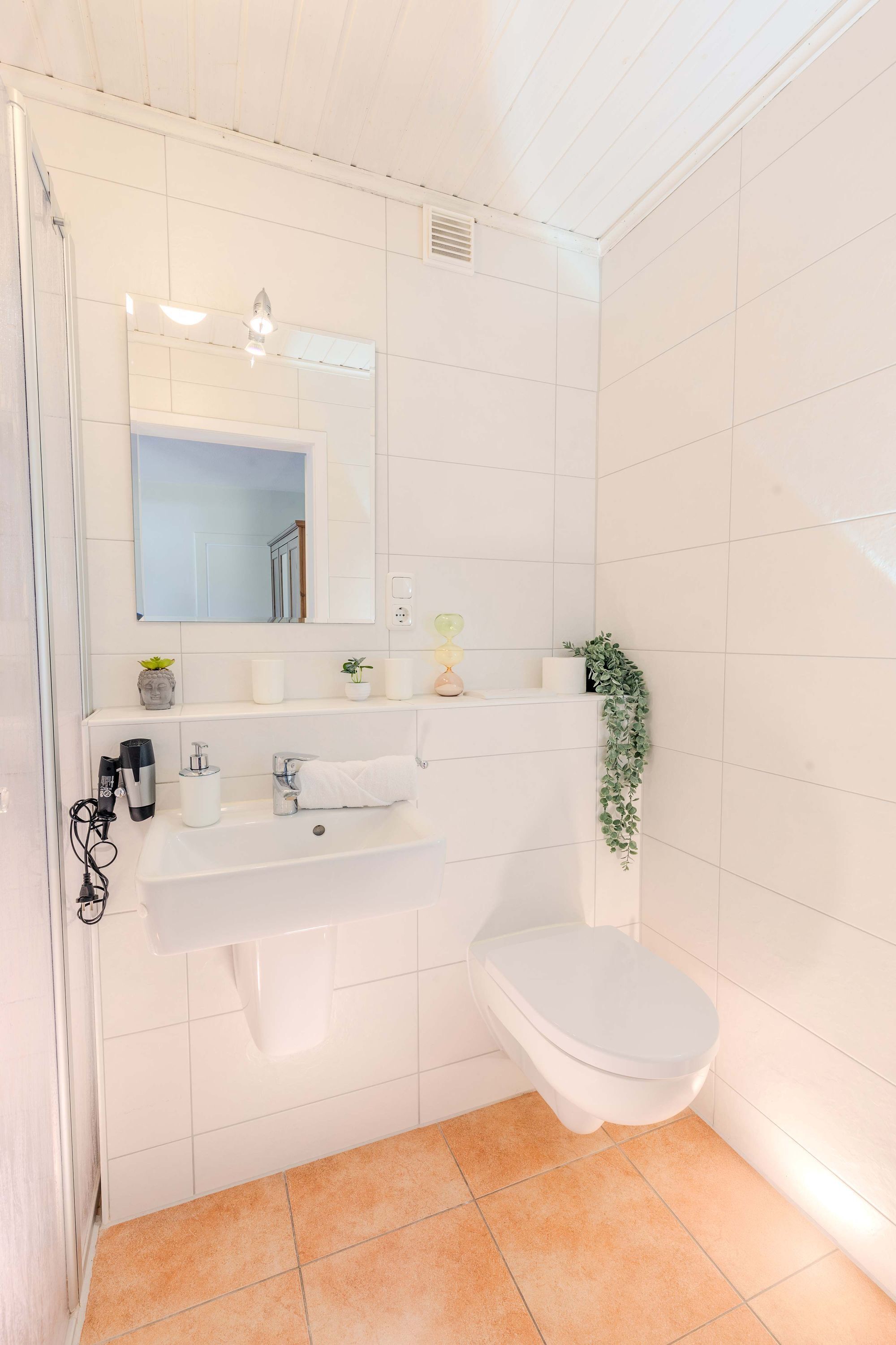 Une salle de bain avec des carreaux blancs, un lavabo blanc, des toilettes, un miroir et une plante.