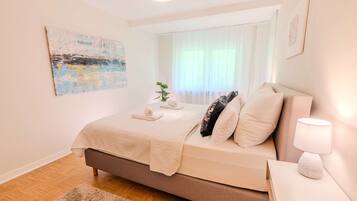 Un dormitorio con cama, lámpara de noche, alfombra, ventana con cortinas y un cuadro en la pared.