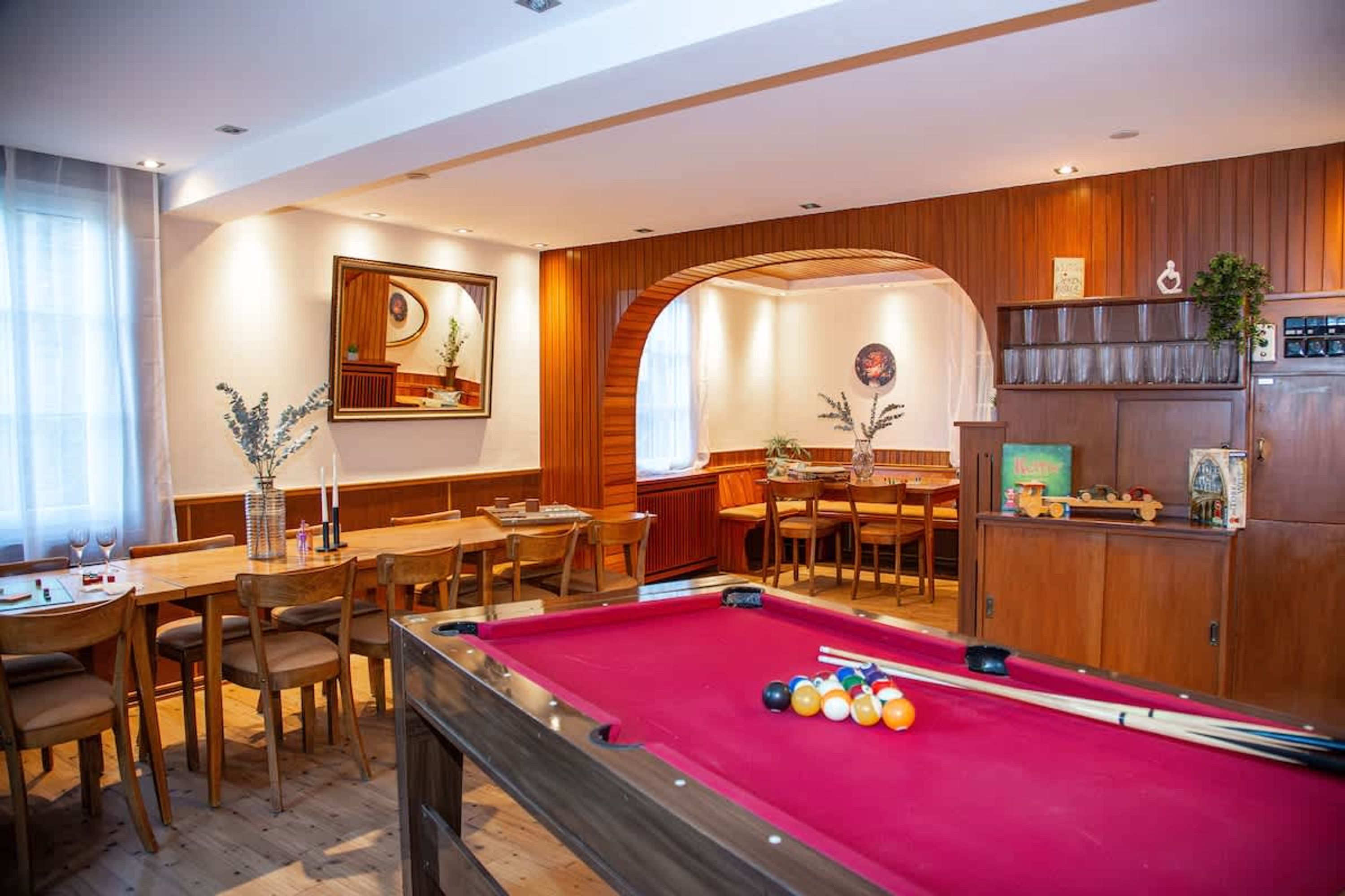 A pool table in a room with wooden paneling, a dining area, and a kitchen area.