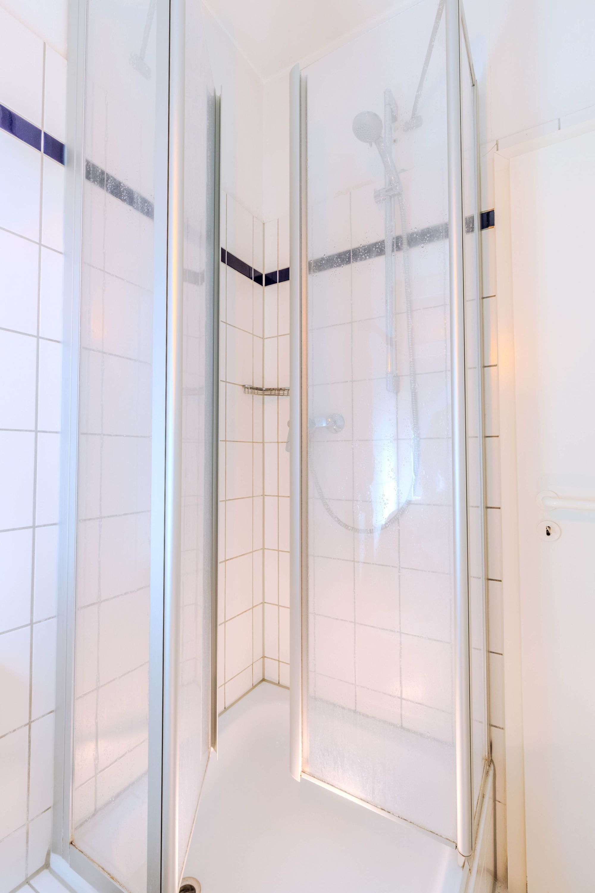 A glass-enclosed shower area with a handheld showerhead, white tiled walls, and a white floor.