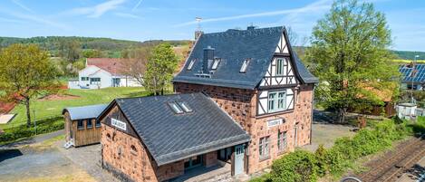 A traditional half-timbered building with a sloped roof, a small adjacent structure, and a railway track nearby.