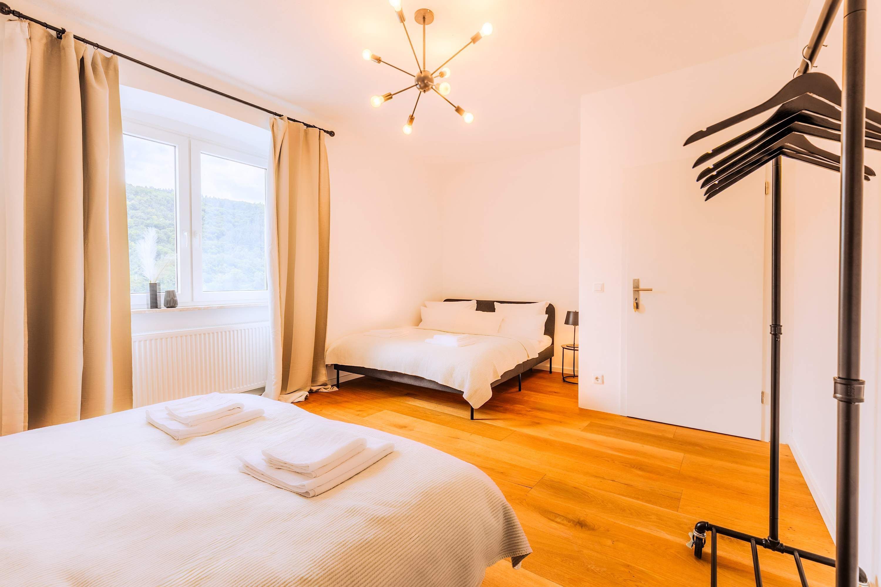 A bedroom with a bed, a window with curtains, a radiator, a ceiling light fixture, and a coat rack.