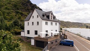 A white building with a steep roof, a blue car parked in front, and a river in the background. - Whirlpool Sauna Kino Villa Klosterschenke (Sankt Goarshausen)