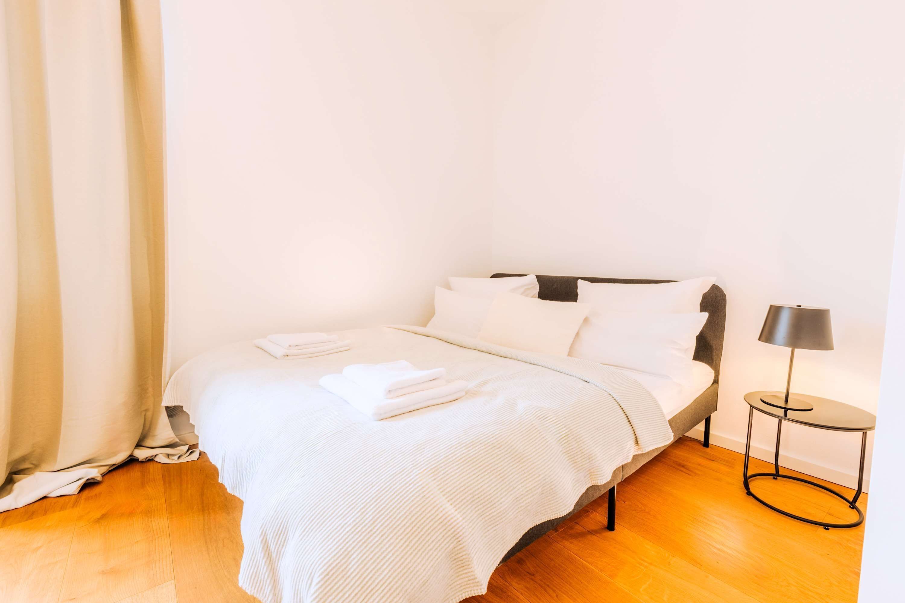 Una cama bien hecha con sábanas blancas, una mesita redonda con lámpara y suelo de madera.