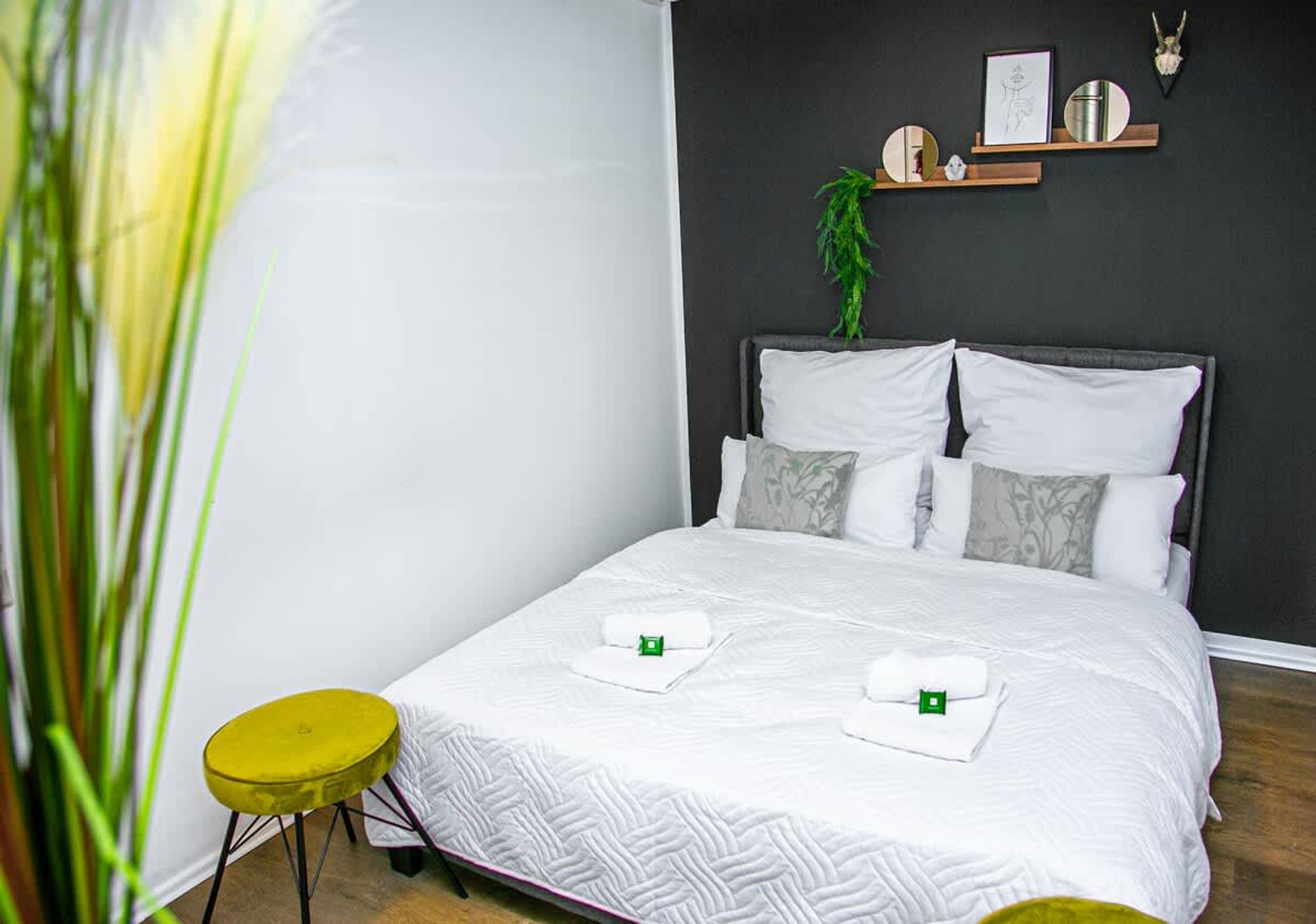 Ein ordentlich bezogenes Bett mit weißer Bettwäsche, eine Zimmerpflanze an der Wand und ein kleiner gelber Hocker.