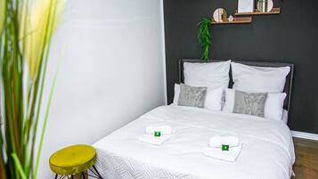 Ein ordentlich bezogenes Bett mit weißer Bettwäsche, eine Zimmerpflanze an der Wand und ein kleiner gelber Hocker.