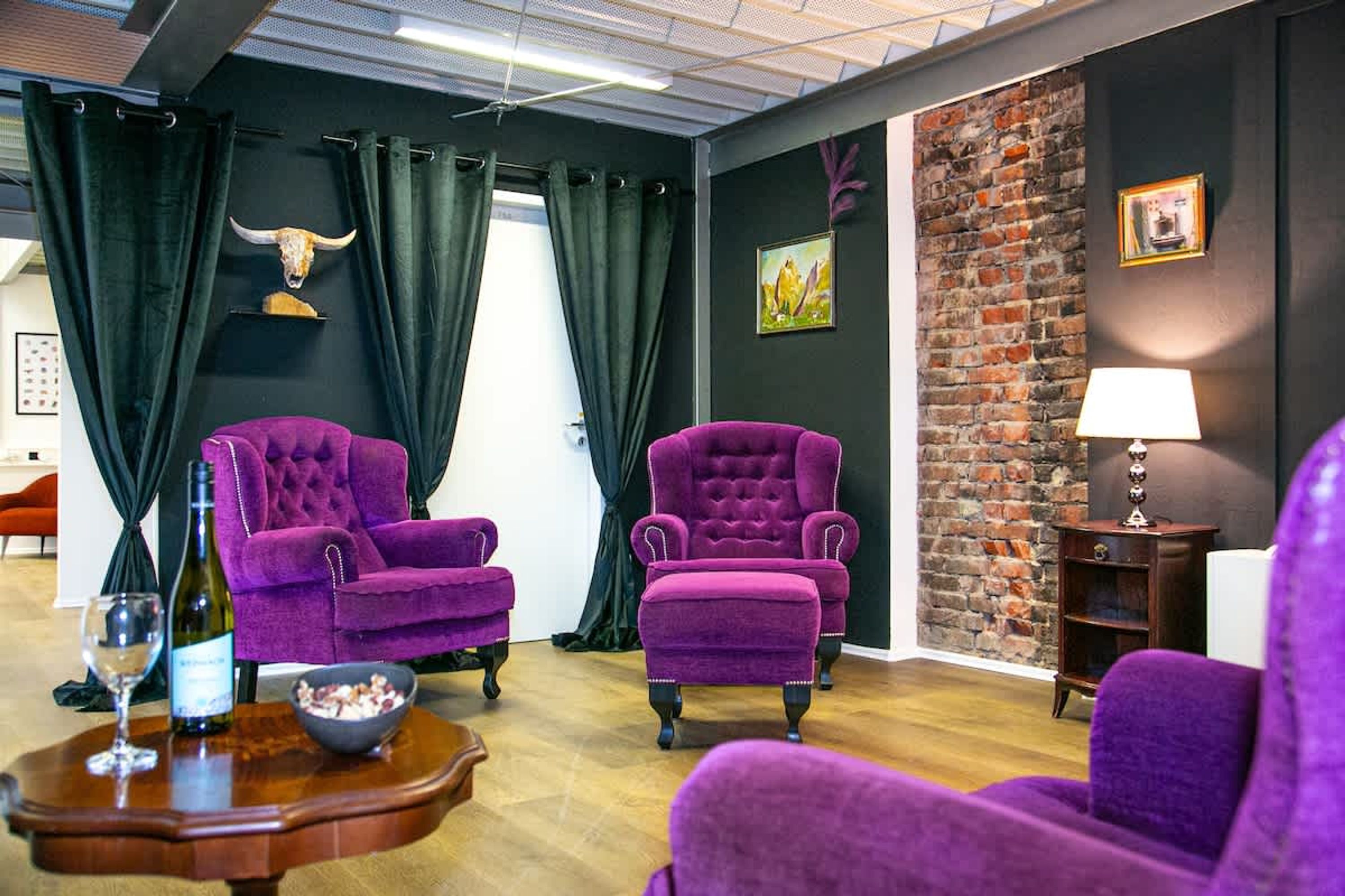 A room with two purple armchairs, a wooden table with a wine bottle and glass, a bowl of snacks, and a brick wall with a picture.