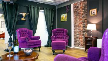 A room with two purple armchairs, a wooden table with a wine bottle and glass, a bowl of snacks, and a brick wall with a picture.