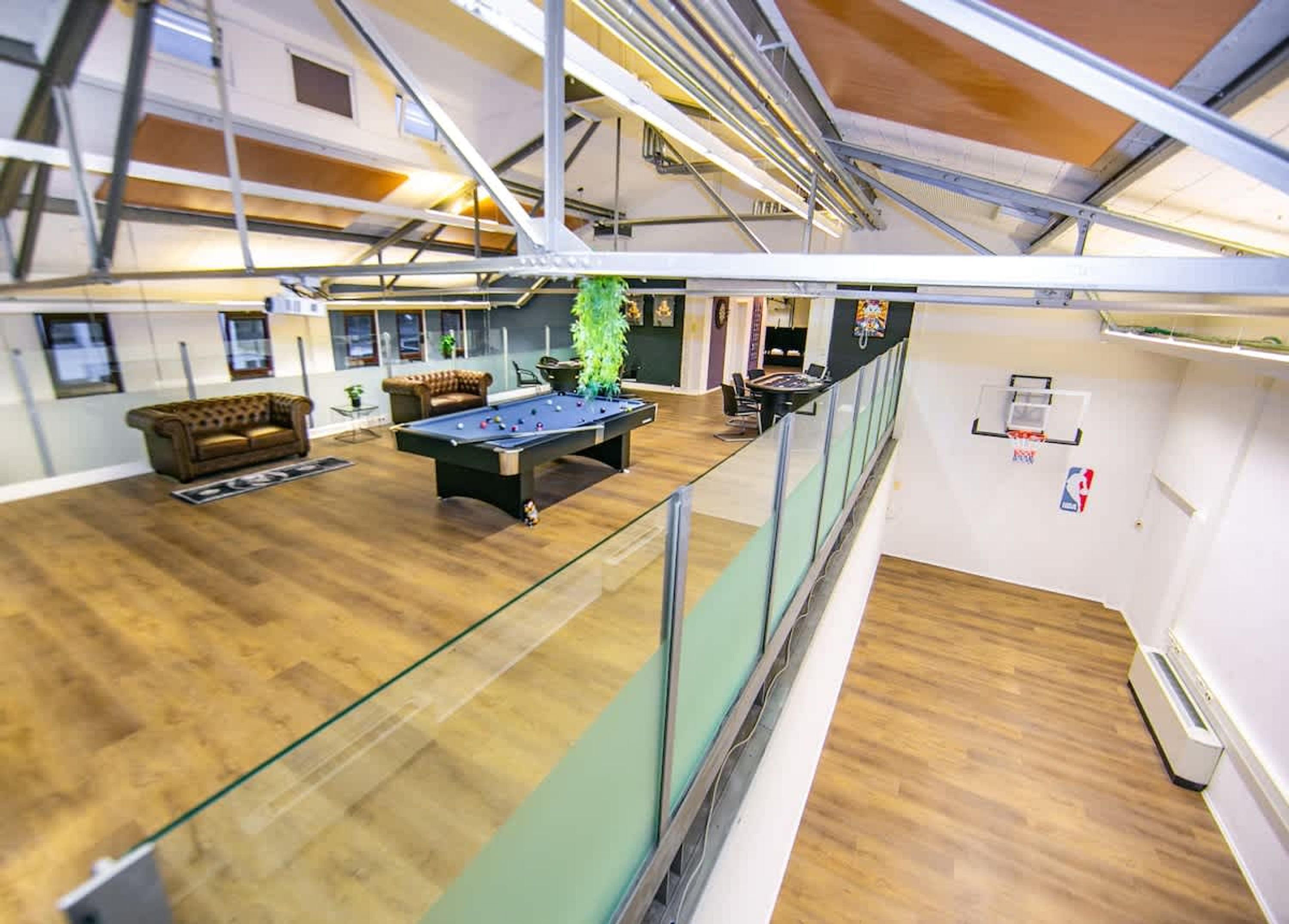 Une table de billard, un canapé et une plante dans une pièce au plancher en bois et dotée d’une rambarde en verre.
