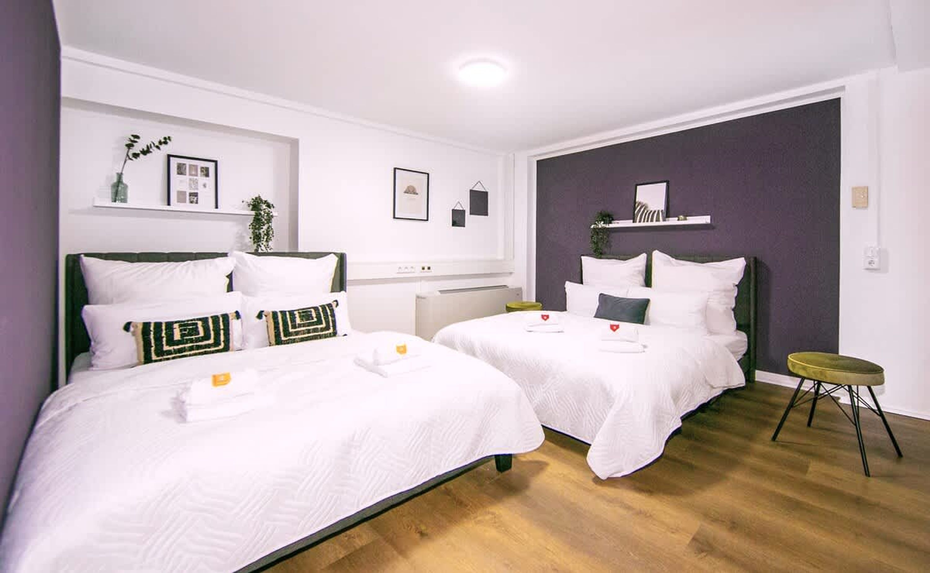 A hotel room with two beds, a small table, a shelf with items, and a plant.