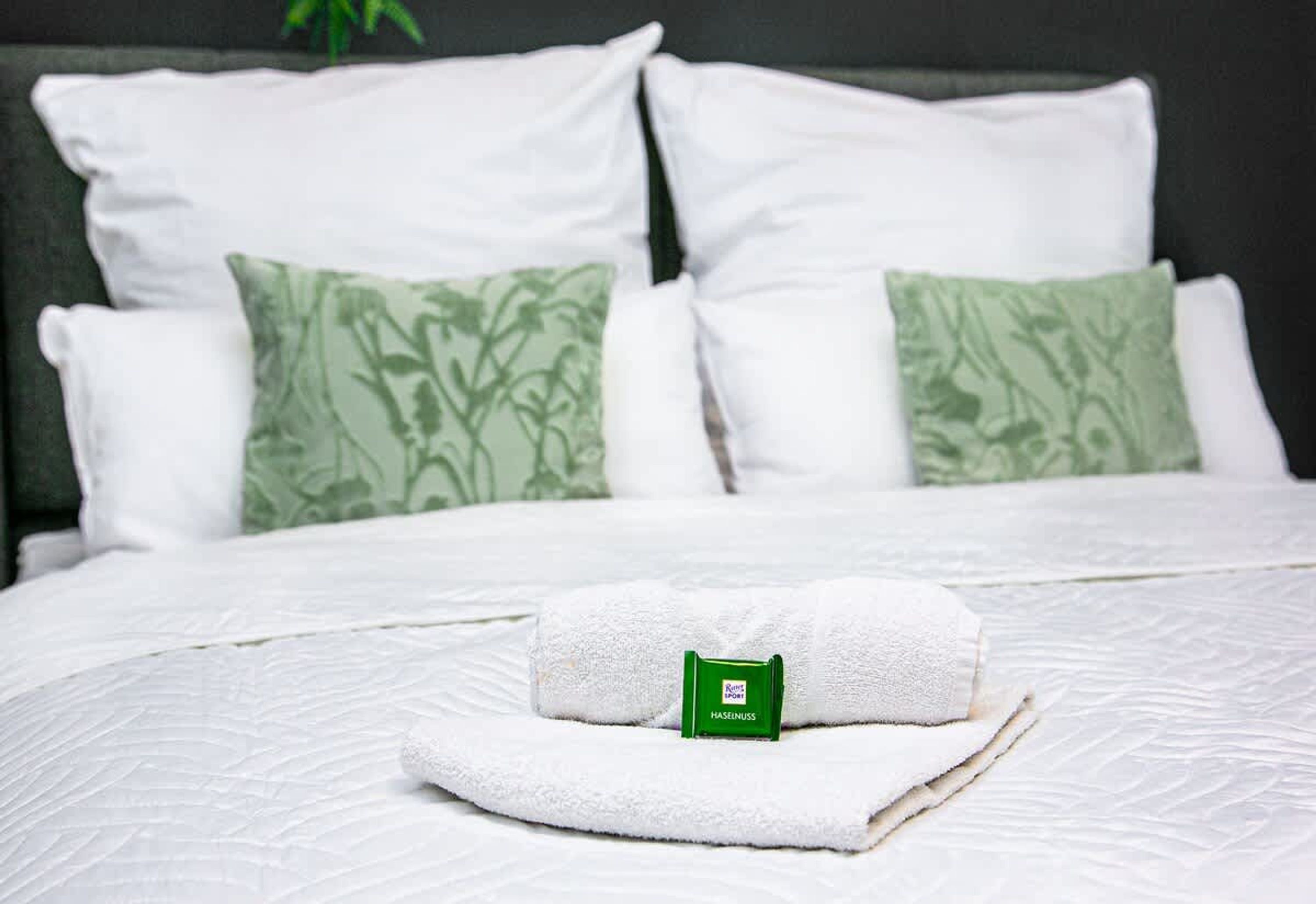Un letto rifatto con lenzuola bianche, un asciugamano arrotolato, una bottiglia verde di Haseenuss e due cuscini con motivo verde.