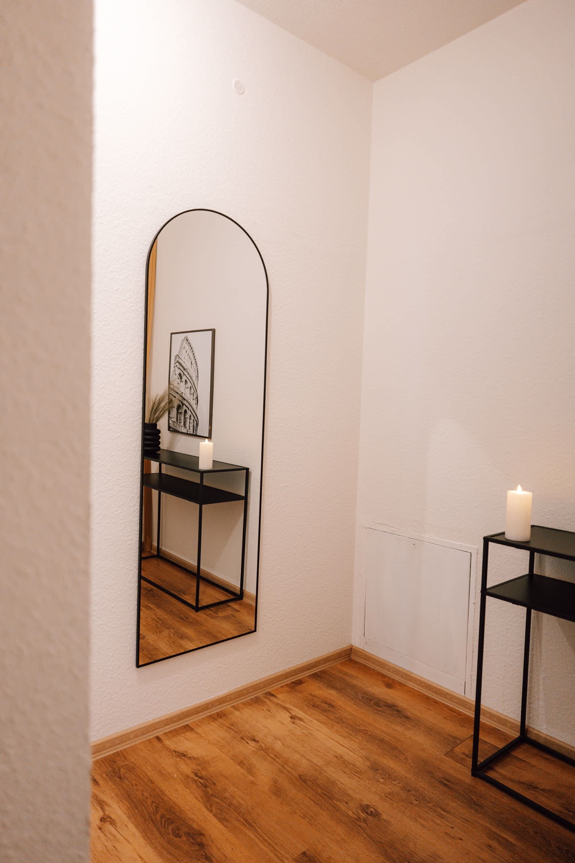 A room with a wooden floor, a black-framed mirror, a small table with a candle, and a framed picture on the wall.
