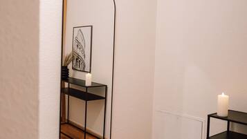 A room with a wooden floor, a black-framed mirror, a small table with a candle, and a framed picture on the wall.