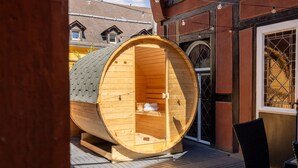 Estructura de sauna de madera con un banco en su interior, ubicada sobre una terraza de madera y una silla cerca.