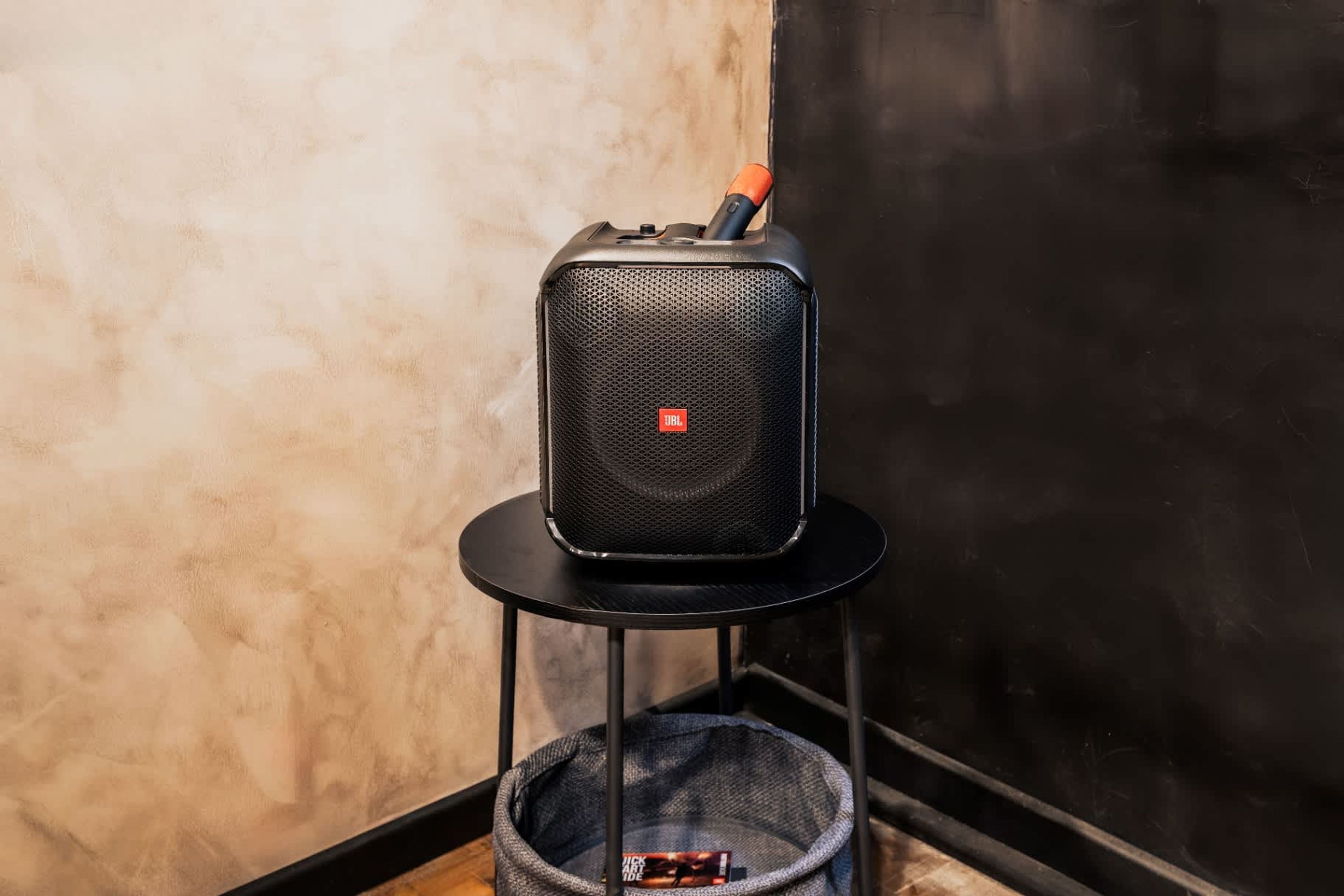 A black JBL speaker on a black stand with a red handle, placed on a textured wall.