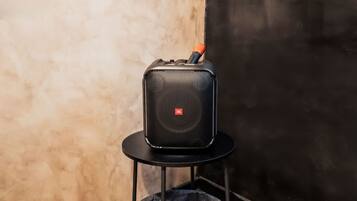 A black JBL speaker on a black stand with a red handle, placed on a textured wall.